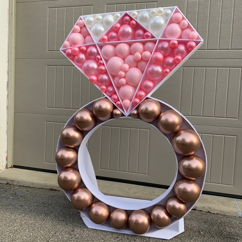 May include: A large white ring made of balloons with a pink and white diamond-shaped balloon structure on top. The ring is made of brown balloons and the diamond is made of pink and white balloons.
