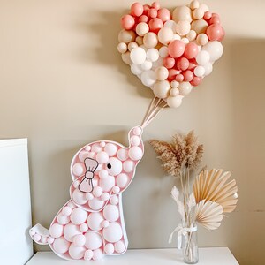 May include: A pink and white balloon elephant with a pink bow, surrounded by a heart-shaped balloon garland in pink and beige.
