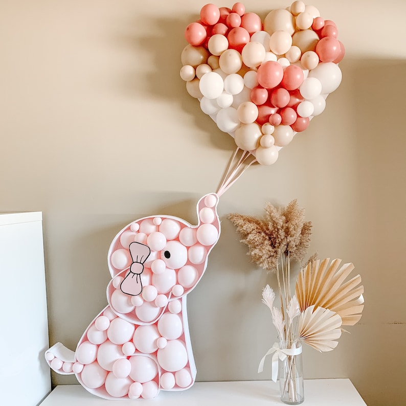 May include: A pink and white balloon elephant with a pink bow, surrounded by a heart-shaped balloon garland in pink and beige.