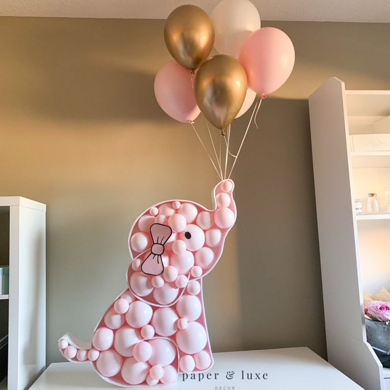 K&ouml;nnte beinhalten: Ein rosa Ballon-Elefant mit einer rosa Schleife, der rosa, wei&szlig;e und goldene Ballons h&auml;lt. Der Elefant besteht aus vielen kleinen Ballons. Der Text "paper & luxe decor" ist unten im Bild sichtbar.