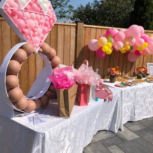May include: A white ring and diamond made of pink and white balloons, with a brown ring of balloons around the base. The balloons are set up on a white table with pink and brown gift bags and a white tablecloth with a floral pattern. There are pink and yellow balloons in the background.