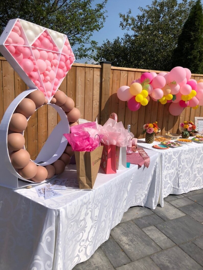 May include: A white ring and diamond made of pink and white balloons, with a brown ring of balloons around the base. The balloons are set up on a white table with pink and brown gift bags and a white tablecloth with a floral pattern. There are pink and yellow balloons in the background.