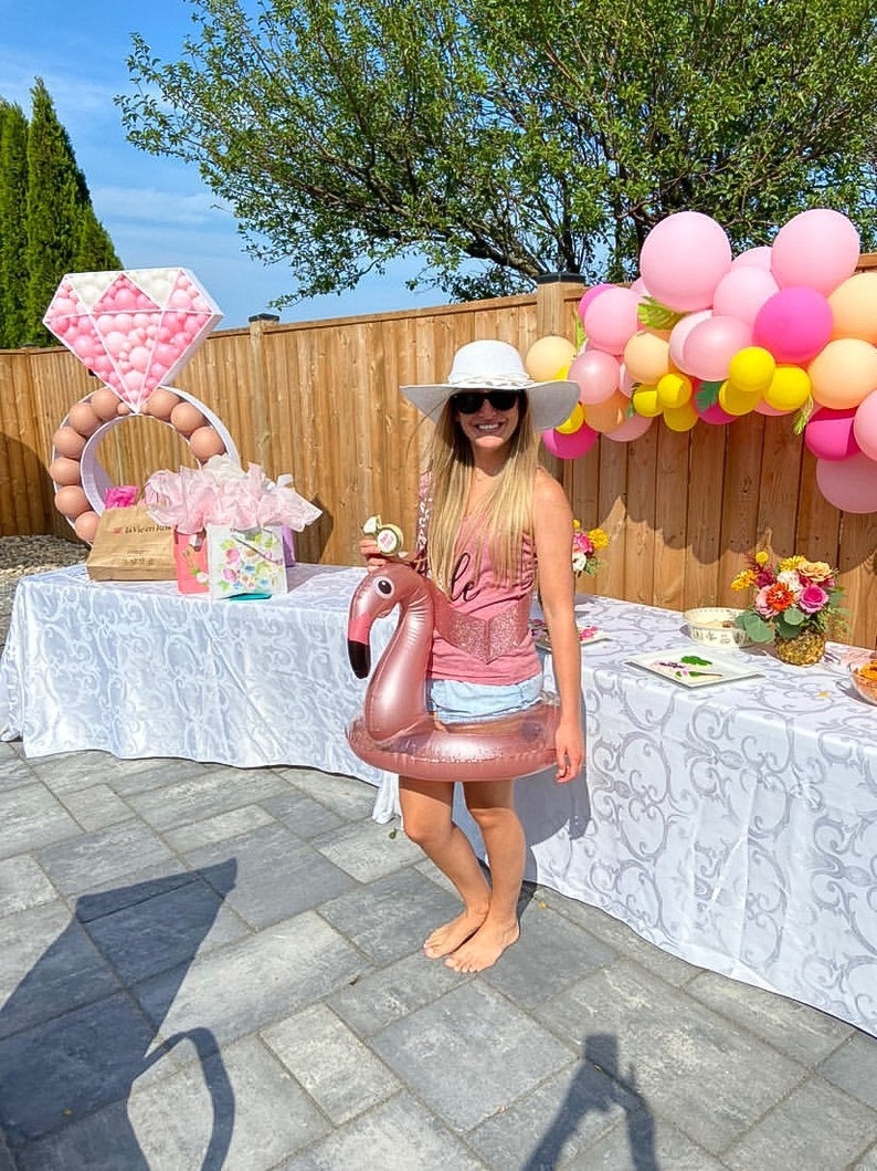 May include: A woman wearing a white hat and a pink tank top with the word "Bride" on it, holds a pink flamingo pool float. The background features a wooden fence with a pink and yellow balloon arch and a large pink diamond ring made of balloons.