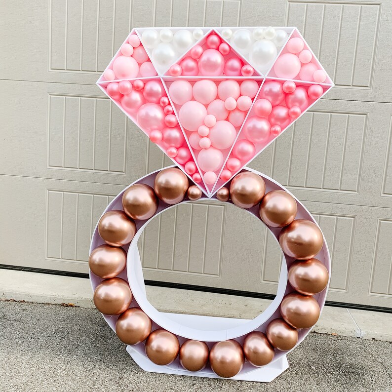May include: A large white ring with a pink and white diamond-shaped balloon decoration. The ring is made of balloons and is decorated with pink and white balloons.