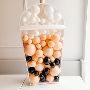 May include: A white balloon sculpture shaped like a boba tea cup with a brown straw. The cup is filled with white, peach, and black balloons.