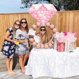 May include: Three women stand in front of a large ring-shaped balloon arch decorated with pink and white balloons. The arch is shaped like a diamond ring. The women are holding signs that say "Team Bride" and "Realls".