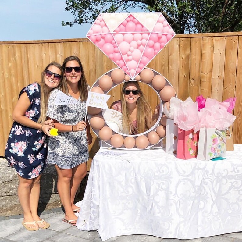 May include: Three women stand in front of a large ring-shaped balloon arch decorated with pink and white balloons. The arch is shaped like a diamond ring. The women are holding signs that say "Team Bride" and "Realls".
