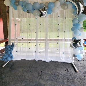 May include: A white sheer curtain backdrop with silver stars and crescent moons printed on it. A blue and silver balloon arch is above the curtain. The balloon arch includes blue, silver, and white balloons. There is a blue balloon elephant in the foreground.