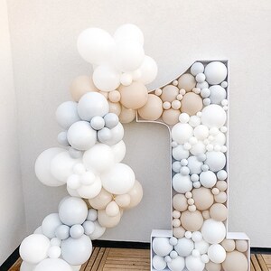K&ouml;nnte beinhalten: Ein gro&szlig;er wei&szlig;er Ballonbogen in Form der Zahl eins mit wei&szlig;en, grauen und beigefarbenen Ballons. Die Ballons sind in einer kaskadierenden Anordnung angeordnet.