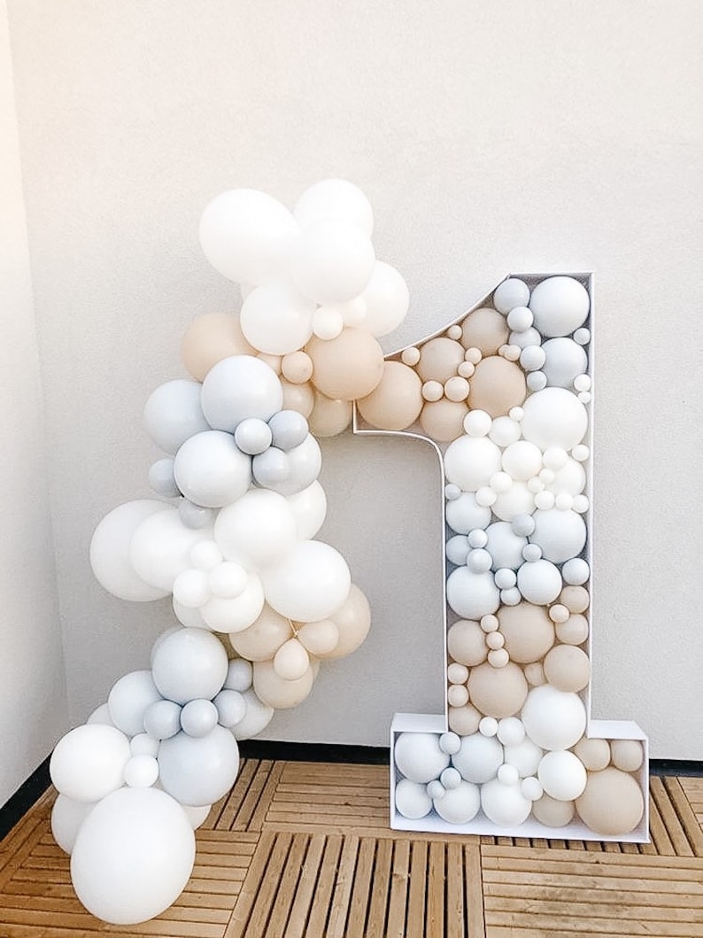 K&ouml;nnte beinhalten: Ein gro&szlig;er wei&szlig;er Ballonbogen in Form der Zahl eins mit wei&szlig;en, grauen und beigefarbenen Ballons. Die Ballons sind in einer kaskadierenden Anordnung angeordnet.