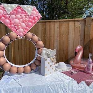 May include: A pink and white balloon ring and diamond shaped balloon decoration for a bridal shower. The ring is made of brown balloons and the diamond is made of pink and white balloons. There is a pink flamingo pool float, a bottle of champagne, and a pink shirt with the word "Bride" on it.