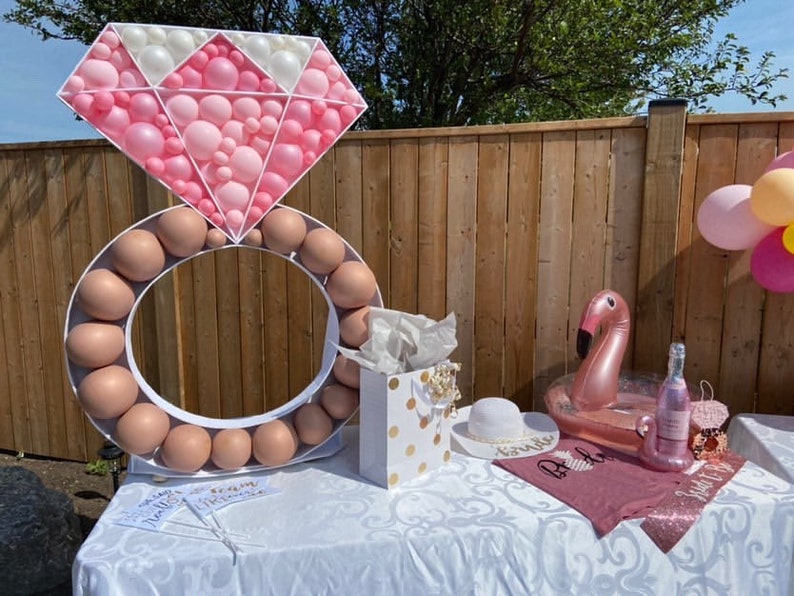 May include: A pink and white balloon ring and diamond shaped balloon decoration for a bridal shower. The ring is made of brown balloons and the diamond is made of pink and white balloons. There is a pink flamingo pool float, a bottle of champagne, and a pink shirt with the word "Bride" on it.