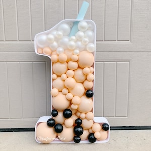 K&ouml;nnte beinhalten: Ein wei&szlig;er Ballon in Form der Nummer eins, der wie ein Boba-Tee-Becher geformt ist. Der Becher ist mit beigefarbenen und wei&szlig;en Ballons gef&uuml;llt, und schwarzen Ballons, die Boba-Perlen darstellen. Ein blauer Strohhalm ragt aus der Oberseite des Bechers. Der Ballon ist mit dem Text "@paperandluxe" verziert.