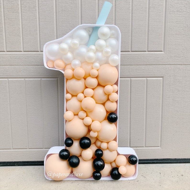 K&ouml;nnte beinhalten: Ein wei&szlig;er Ballon in Form der Nummer eins, der wie ein Boba-Tee-Becher geformt ist. Der Becher ist mit beigefarbenen und wei&szlig;en Ballons gef&uuml;llt, und schwarzen Ballons, die Boba-Perlen darstellen. Ein blauer Strohhalm ragt aus der Oberseite des Bechers. Der Ballon ist mit dem Text "@paperandluxe" verziert.
