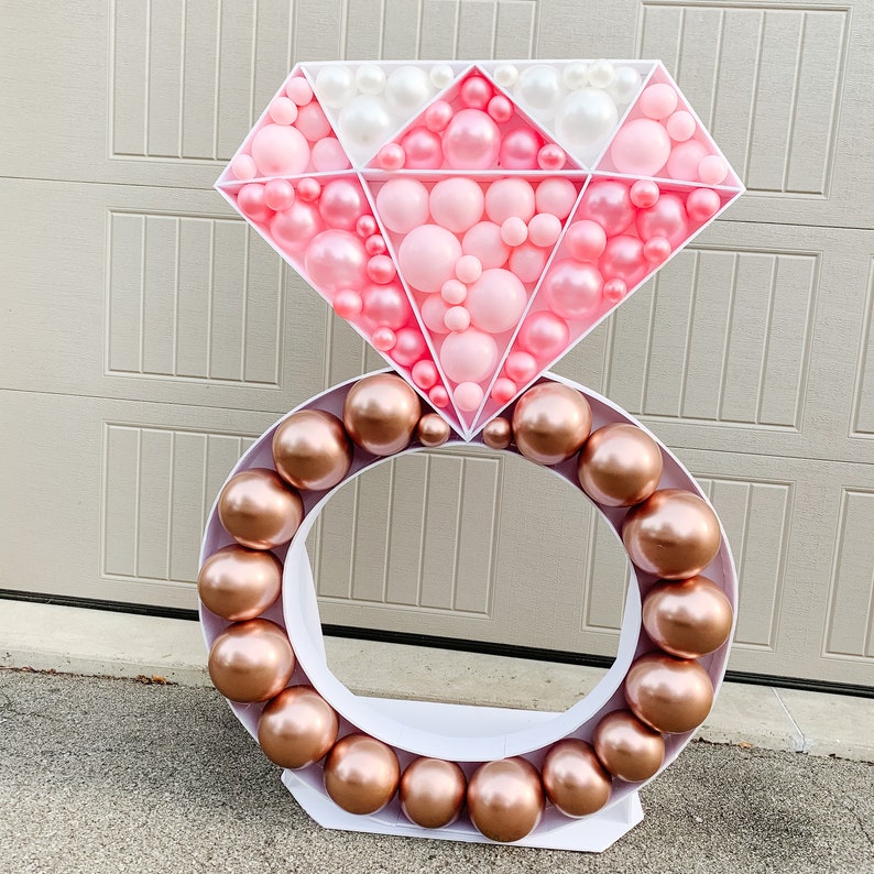 May include: A large white diamond-shaped balloon arch made of pink and white balloons. The arch is attached to a white ring made of brown balloons.