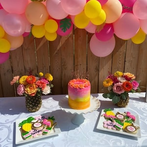 May include: A pink and yellow balloon arch with green leaves hangs above a table with a pink and yellow ombre cake, pineapple centerpieces, and decorated cookies. The cookies feature pink flamingos, pineapples, and the text "Let's Flamingo".