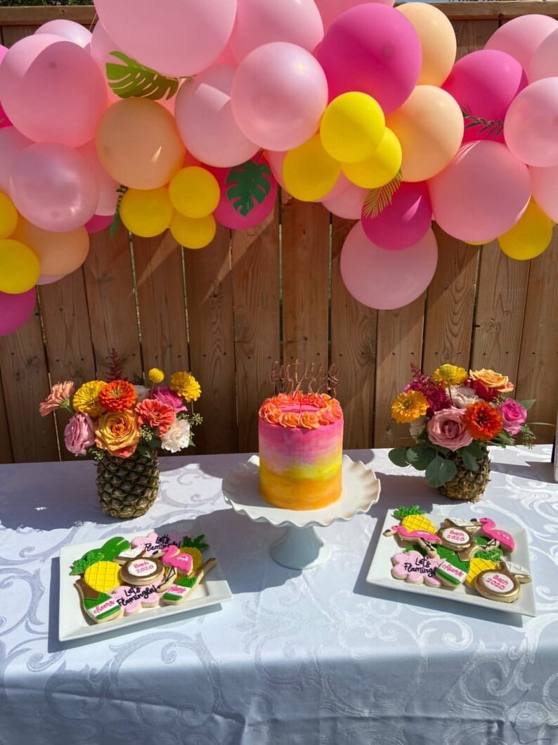 May include: A pink and yellow balloon arch with green leaves hangs above a table with a pink and yellow ombre cake, pineapple centerpieces, and decorated cookies. The cookies feature pink flamingos, pineapples, and the text "Let's Flamingo".