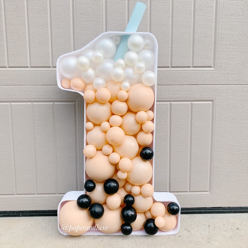 K&ouml;nnte beinhalten: Ein wei&szlig;er Ballon in Form der Nummer eins, der wie ein Boba-Tee-Becher geformt ist. Der Becher ist mit beigefarbenen und wei&szlig;en Ballons gef&uuml;llt, und schwarzen Ballons, die Boba-Perlen darstellen. Ein blauer Strohhalm ragt aus der Oberseite des Bechers. Der Ballon ist mit dem Text "@paperandluxe" verziert.
