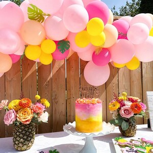 May include: A pink and yellow balloon arch with green tropical leaves, a pink and yellow ombre cake with a gold topper that says "Summer Vibes", and two pineapple vases with pink and orange flowers. The cake is on a white cake stand and the flowers are in two pineapple vases. There are also cookies on a white plate with the text "Let's Flamingo!"