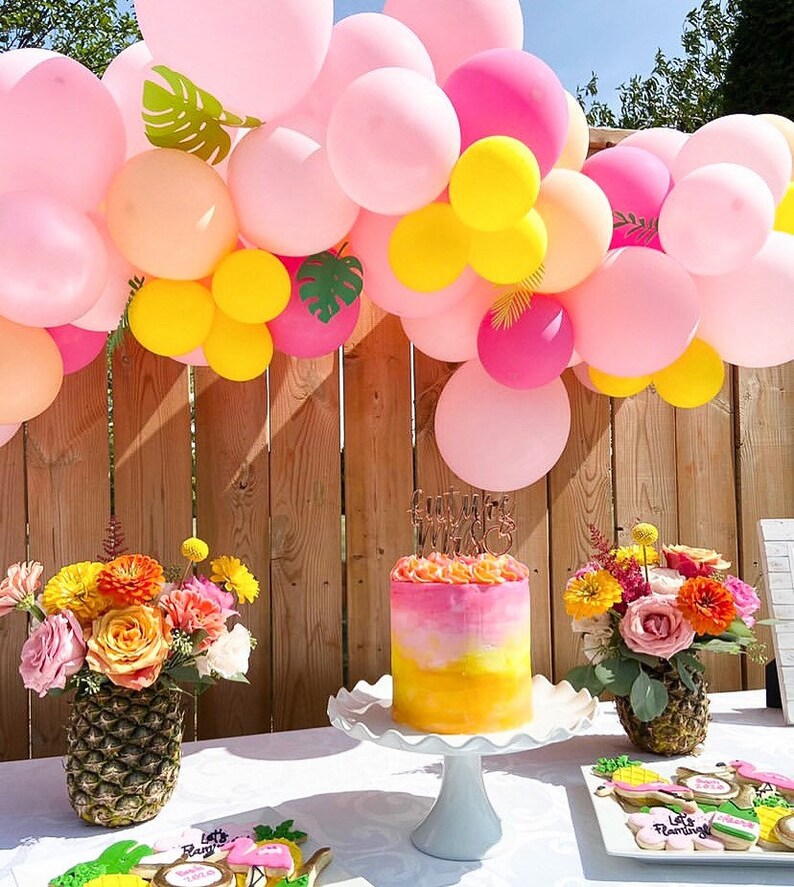May include: A pink and yellow balloon arch with green tropical leaves, a pink and yellow ombre cake with a gold topper that says "Summer Vibes", and two pineapple vases with pink and orange flowers. The cake is on a white cake stand and the flowers are in two pineapple vases. There are also cookies on a white plate with the text "Let's Flamingo!"
