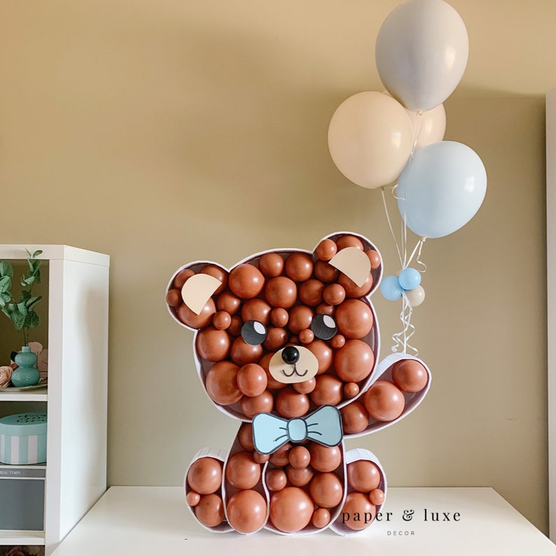 K&ouml;nnte beinhalten: Ein brauner Ballon-B&auml;r mit einer blauen Schleife, der einen Strau&szlig; Ballons in Blau, Beige und Grau h&auml;lt. Die Ballons sind mit einem wei&szlig;en Band zusammengebunden. Der B&auml;r sitzt auf einer wei&szlig;en Oberfl&auml;che mit dem Text "paper & luxe decor" darunter.