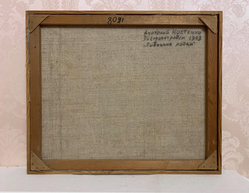 May include: The back of a framed painting on canvas. The canvas is a light beige color and the frame is a light brown wood. The painting is signed "Anatoliy Kostenko Dnepropetrovsk 1993 "Rybachie lodki" ".