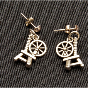 May include: Silver-toned earrings featuring a miniature spinning wheel design. The earrings have a post-back closure and a detailed representation of a spinning wheel, with a wheel and frame. The earrings are displayed on a dark textured surface.