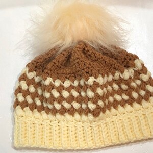 May include: A crocheted beanie hat with a brown and white checkered pattern and a fluffy white pom pom on top.