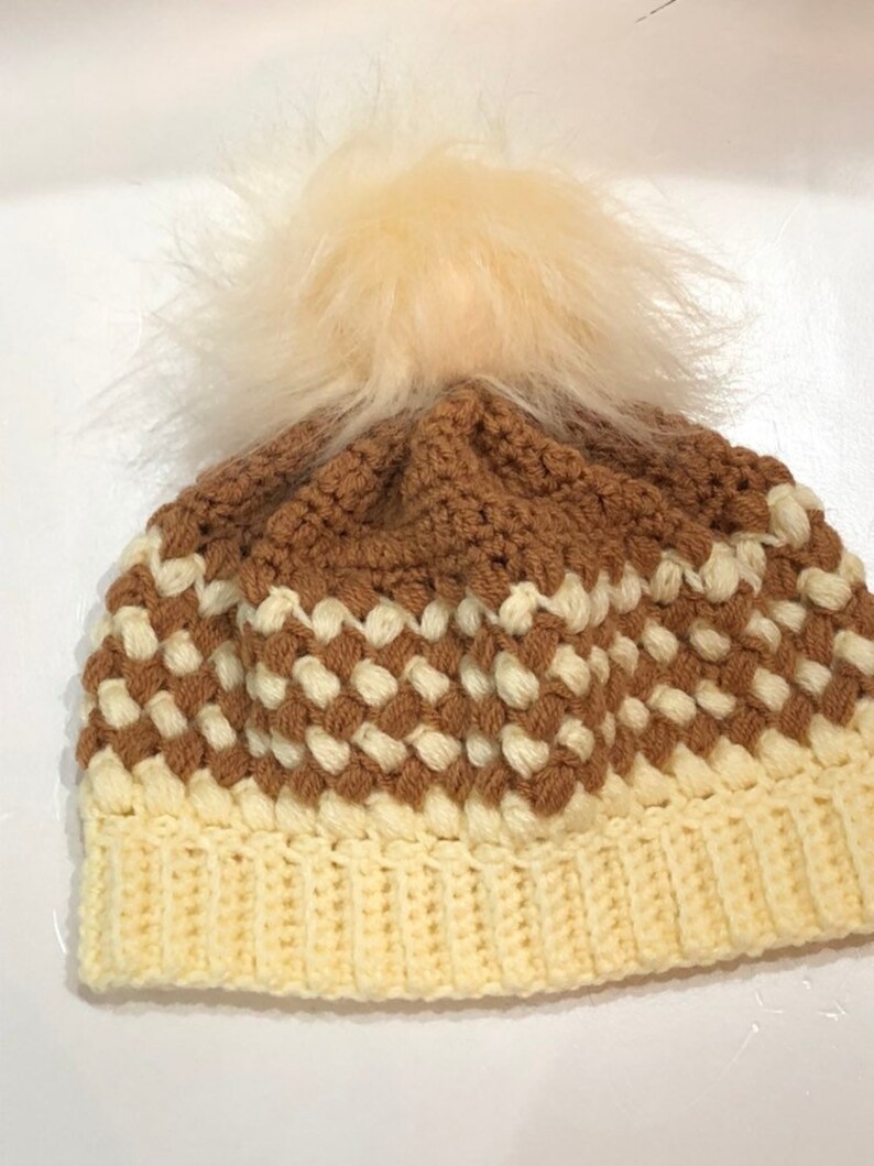 May include: A crocheted beanie hat with a brown and white checkered pattern and a fluffy white pom pom on top.
