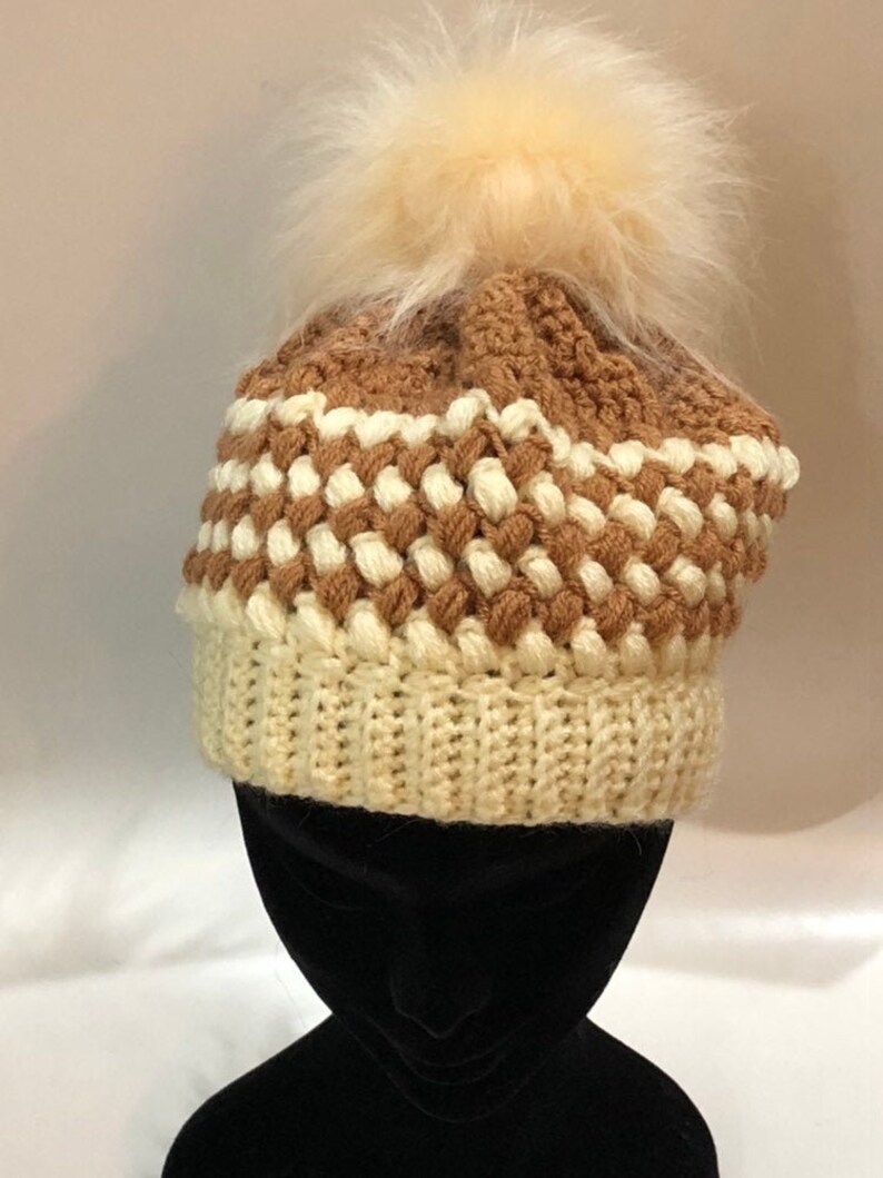 May include: A brown and white crocheted beanie with a fluffy white pom pom. The beanie has a textured pattern and is made with soft yarn.
