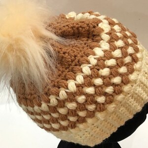 May include: A brown and white crocheted beanie with a fluffy white pom pom.