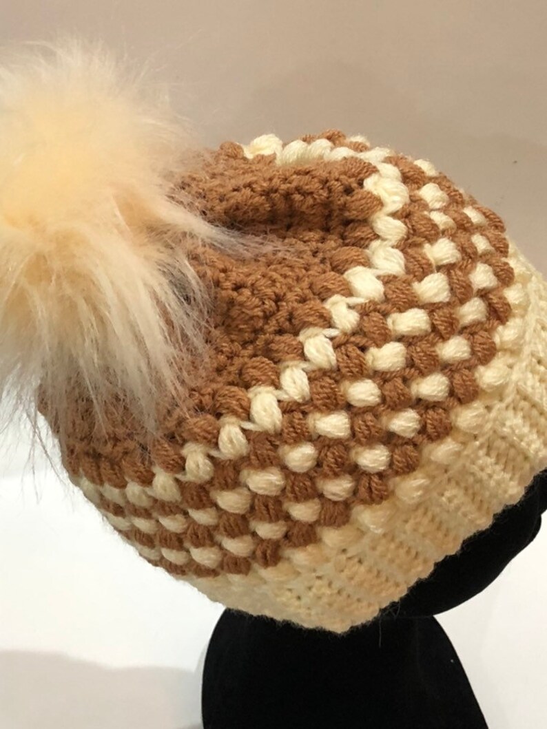 May include: A brown and white crocheted beanie with a fluffy white pom pom.