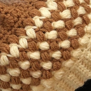 May include: Close-up of a brown and white crocheted hat with a textured pattern. The hat is made with two different shades of yarn, creating a striped effect.
