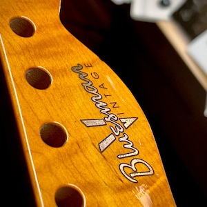 May include: Close-up of a vintage, light brown guitar neck with a dark brown, engraved logo that reads "Bluesman Vintage".