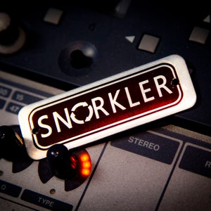 May include: A silver metal label with the word "SNORKLER" in red letters on a black background. The label is lying on top of a silver and black audio mixing board.