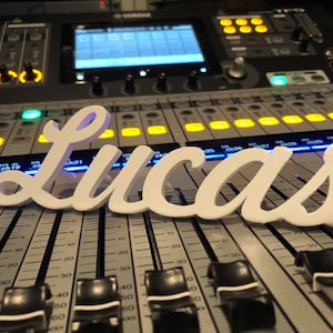 Op de afbeelding: Witte plastic letters die de naam "Lucas" spellen op een zilveren mengpaneel met zwarte knoppen en faders.