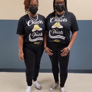 May include: Two people wearing black t-shirts with the text "Chucks & Pearls 2021" and gold glitter accents. The shirts feature a pair of yellow sneakers with gold glitter accents. Both people are wearing black pants and white sneakers.