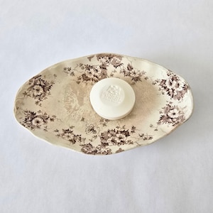 May include: Oval-shaped soap dish with a floral pattern in brown and cream. A round, white bar of soap rests in the center of the dish. The soap dish has a slightly irregular edge.