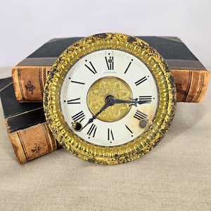 May include: Antique clock face with a gold-colored, ornate bezel. The white clock face features black Roman numerals and black clock hands. The center of the clock face has a decorative gold design. The clock face is labeled "Sessions".