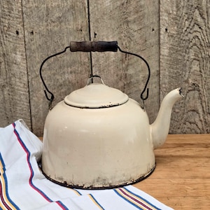 May include: Cream-colored vintage enamelware kettle with a black rim and handle. The kettle has a wooden handle and a spout. The kettle is sitting on a wooden surface with a striped cloth in the foreground.