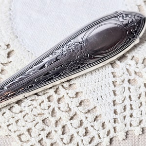 May include: Close-up of a silver spoon with intricate floral and scrollwork detailing. The spoon features an oval design at the top and the word "STERLING" is visible. The spoon is set against a white lace background.