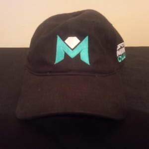 May include: Black baseball cap with a teal and white logo. The logo is a stylized "M" with a diamond shape in the center. The cap also has text on the side in white and teal.