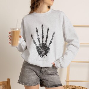 Pode incluir: Uma camisola de gola redonda cinzenta clara com um gráfico preto de uma impressão de mão esquelética centrada no peito. Abaixo do gráfico, o texto "WE ARE NOT ALONE" está impresso em letras maiúsculas pretas pequenas. Mangas compridas.