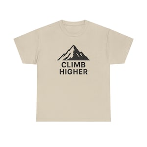 May include: A beige t-shirt featuring a black mountain graphic and the words "CLIMB HIGHER" in a bold, sans-serif font. The design is centered on the front of the shirt, creating a simple yet motivational aesthetic.