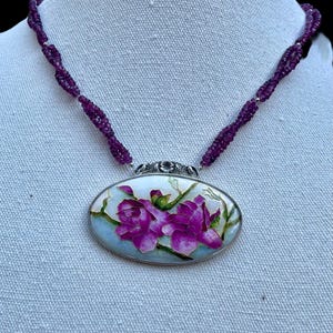 May include: A necklace featuring a pendant with a floral design. The oval pendant has a silver-colored frame and displays a painting of purple flowers. The necklace has a beaded strand of purple beads.