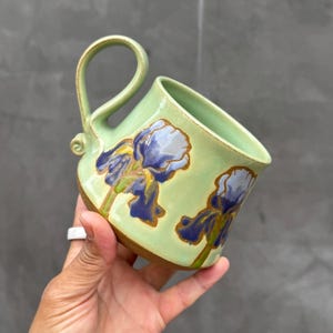 May include: A light green ceramic mug featuring a hand-painted blue and yellow iris flower design. The mug has a tall, curved handle with a small decorative swirl at the base and a contrasting unglazed brown clay bottom edge.
