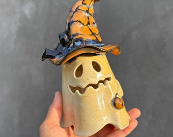 May include: A ceramic ghost figurine featuring a glossy, orange and black glazed witch hat. The ghost has a smooth, light beige body with cut-out eyes and a jagged mouth, and a small orange pumpkin charm attached to its side.