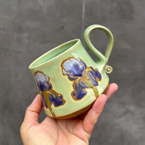 Può includere: Una tazza in ceramica verde chiaro con fiori di iris dipinti a mano in viola e blu con contorni marroni. La tazza presenta una finitura liscia e lucida, un manico curvo con una piccola spirale decorativa alla base e un fondo in argilla marrone.