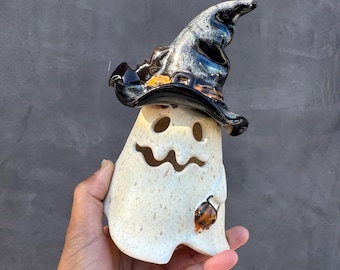 May include: A ceramic ghost figurine with a speckled cream-colored body, featuring cutouts for eyes and a mouth. The ghost wears a glossy, dark, textured witch hat with orange accents and holds a small orange pumpkin in one hand.