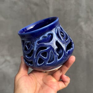 May include: A handcrafted, cobalt blue ceramic vase with a unique openwork design. The vase features intricate cut-out patterns, creating a decorative and artistic look. The interior is a darker blue, and the rim has a lighter blue glaze.